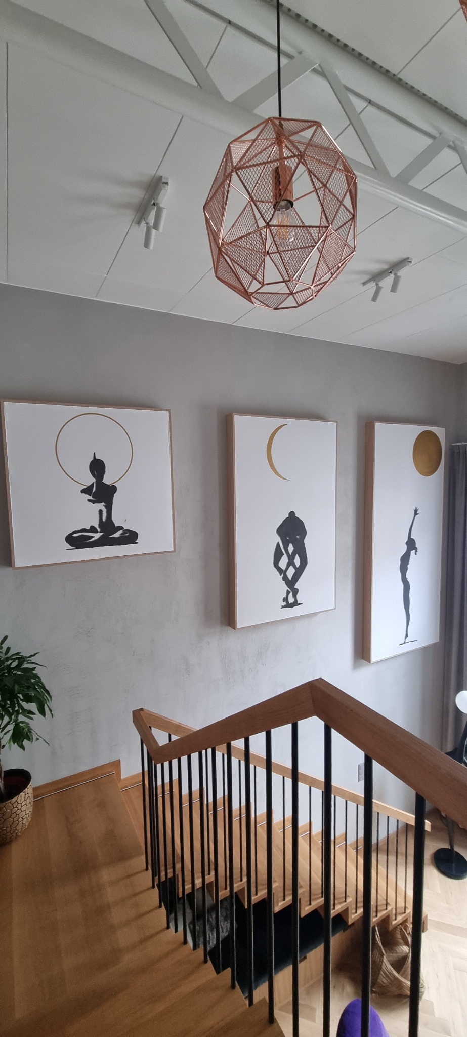 Trzy abstrakcyjne obrazy z motywami jogi i astrologii wiszące na szarej ścianie nad drewnianymi schodami z czarnymi balustradami, oświetlone miedzianą lampą wiszącą o geometrycznym kształcie.