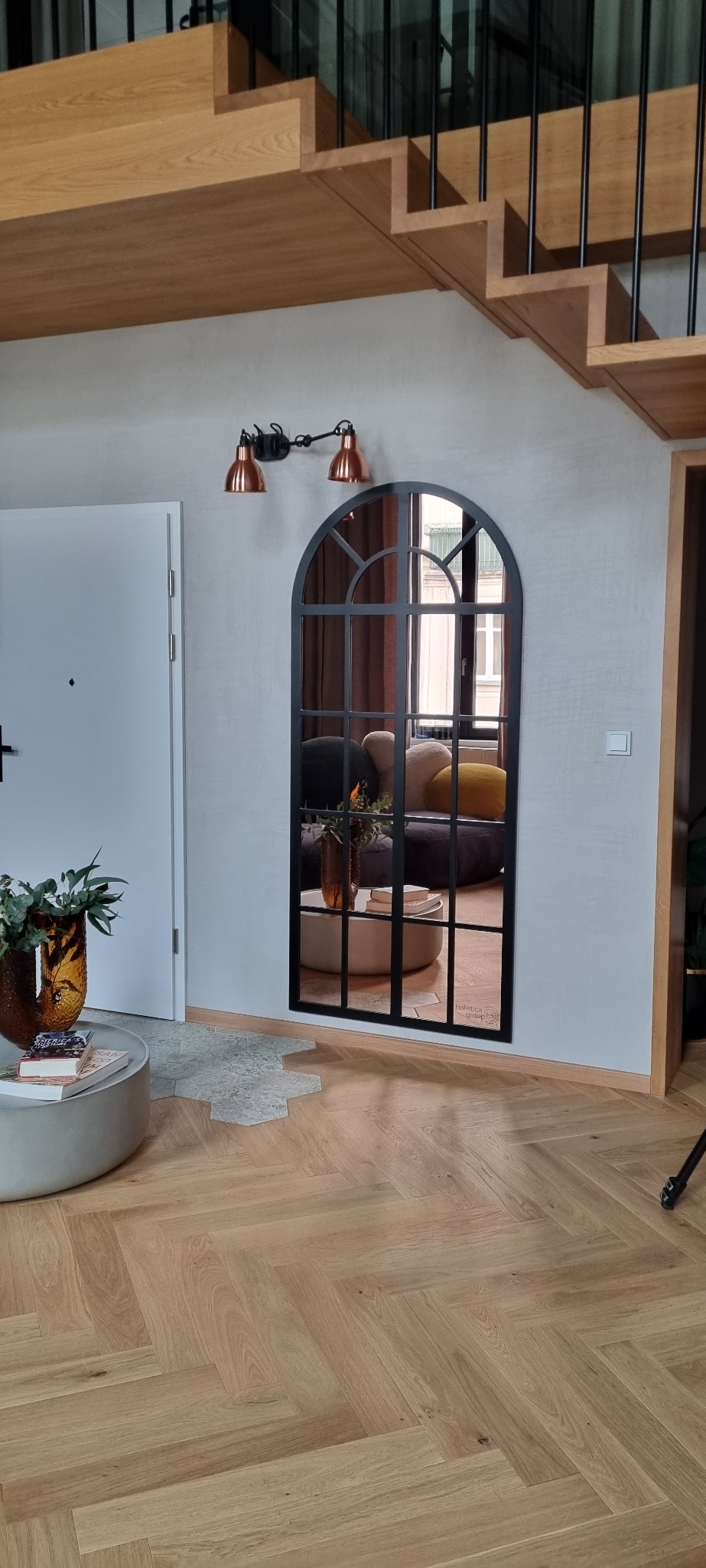 Aranżacja wnętrza z dużym lustrem w czarnej ramie, odbijającym część pokoju z okrągłym stolikiem i pufami, pod schodami z drewnianymi stopniami i czarnymi balustradami, na ścianie lampki w stylu...