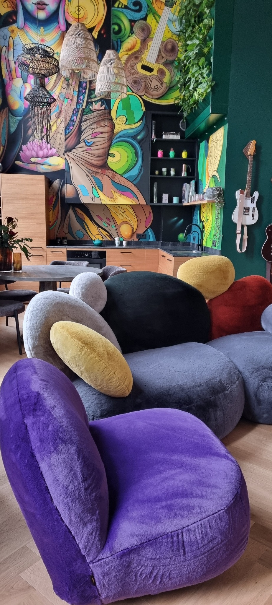 Wnętrze pokoju z kolorowym muralem na ścianie, designerskimi lampami i sofą modułową z pufami w kształcie kamieni w różnych kolorach oraz fotelem w kolorze fioletowym.