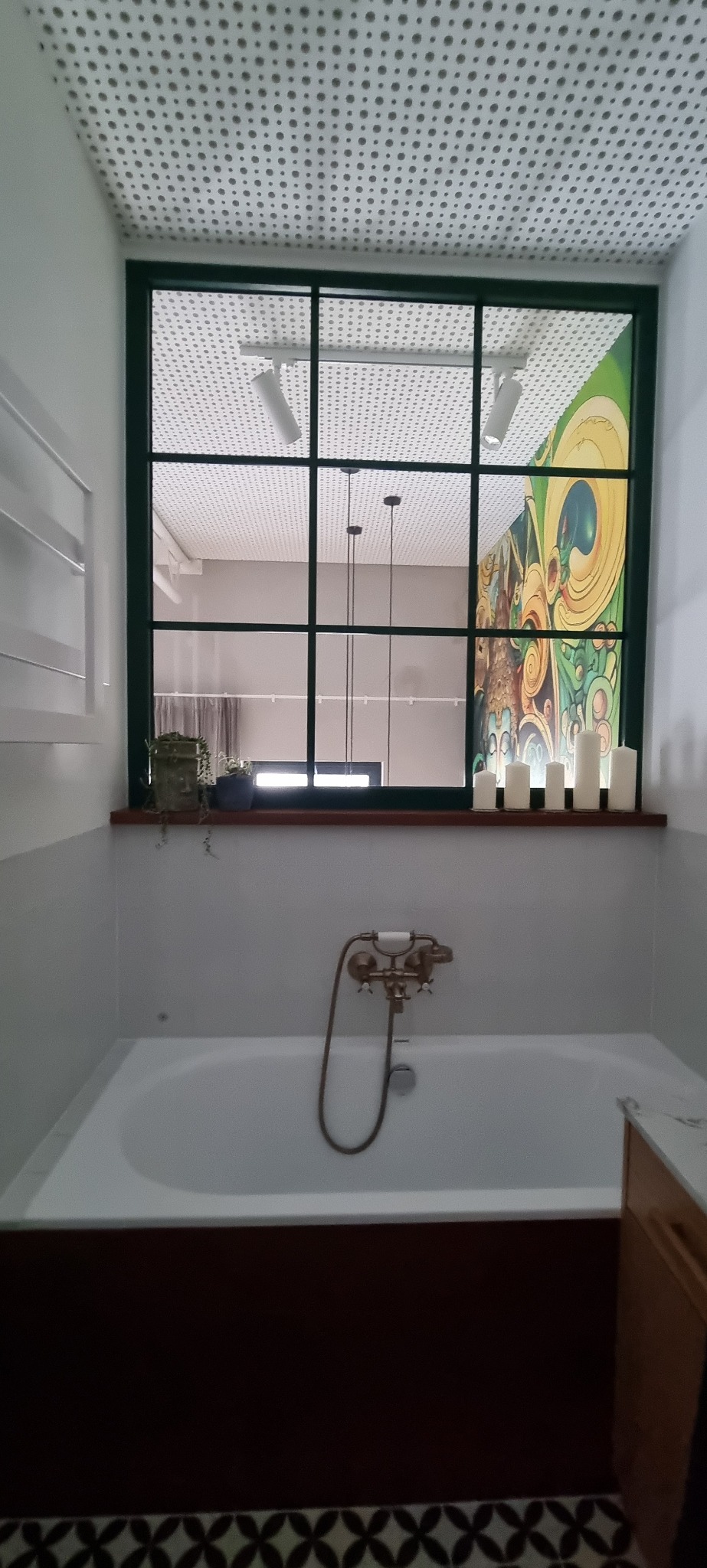 Biała wanna z ciemnobrązowym obramowaniem, bateria w stylu retro i dekoracyjne świece na parapecie okna z zieloną ramą, widok na sufit z okrągłymi otworami i fragment kolorowej grafiki na ścianie.