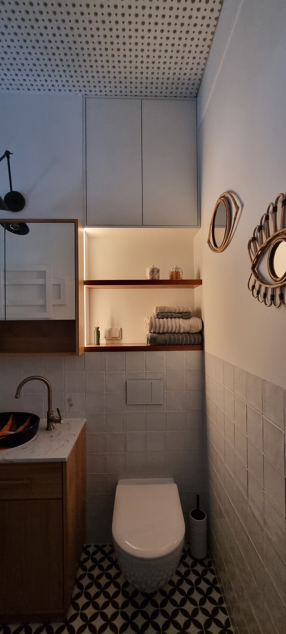 Nowoczesna łazienka z geometryczną podłogą, drewnianą szafką pod umywalką z marmurowym blatem, toaletą, półką z podświetleniem LED i dekoracyjnymi lustrami w kształcie oka.