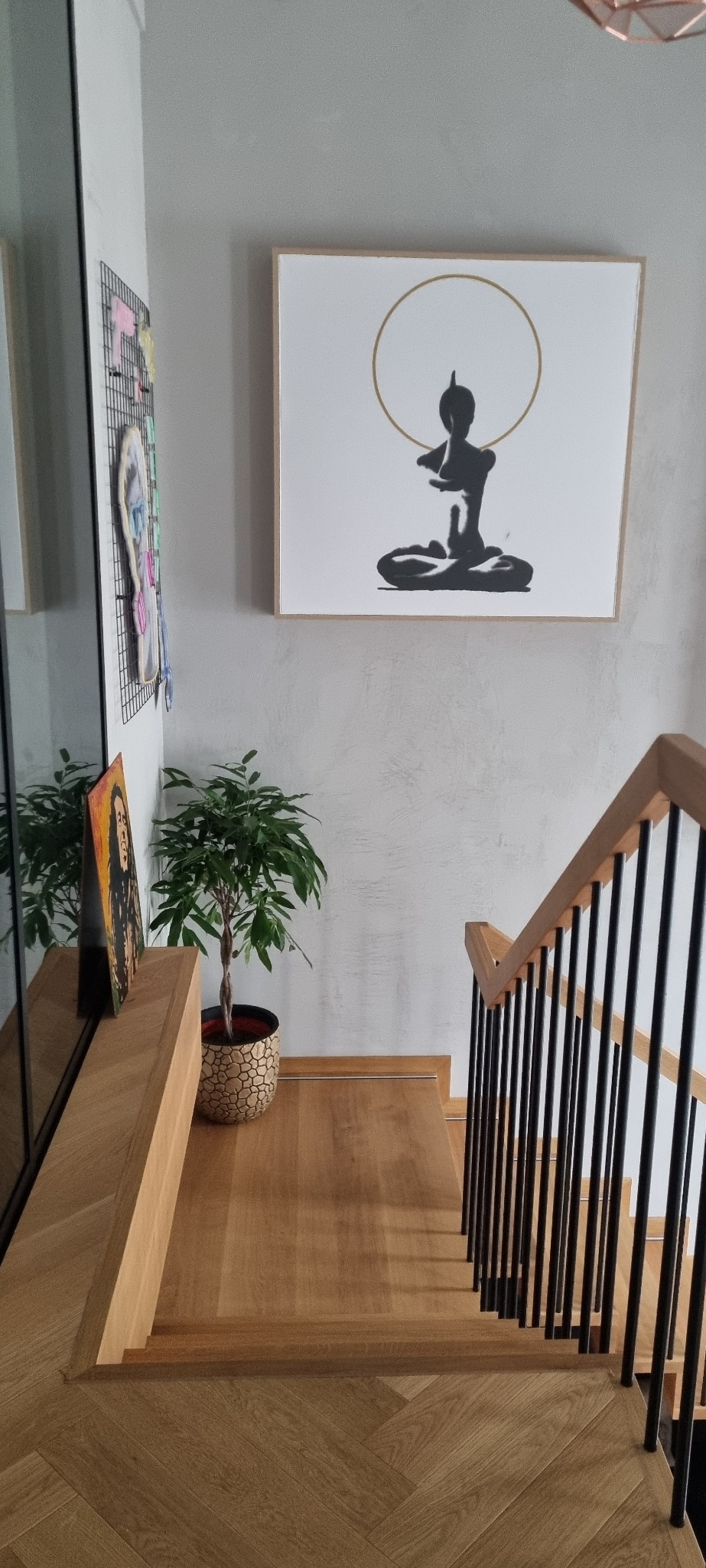 Schody z drewnianymi stopniami ułożonymi w jodełkę, czarne metalowe balustrady, na ścianie obraz z motywem buddyjskim i roślina doniczkowa.