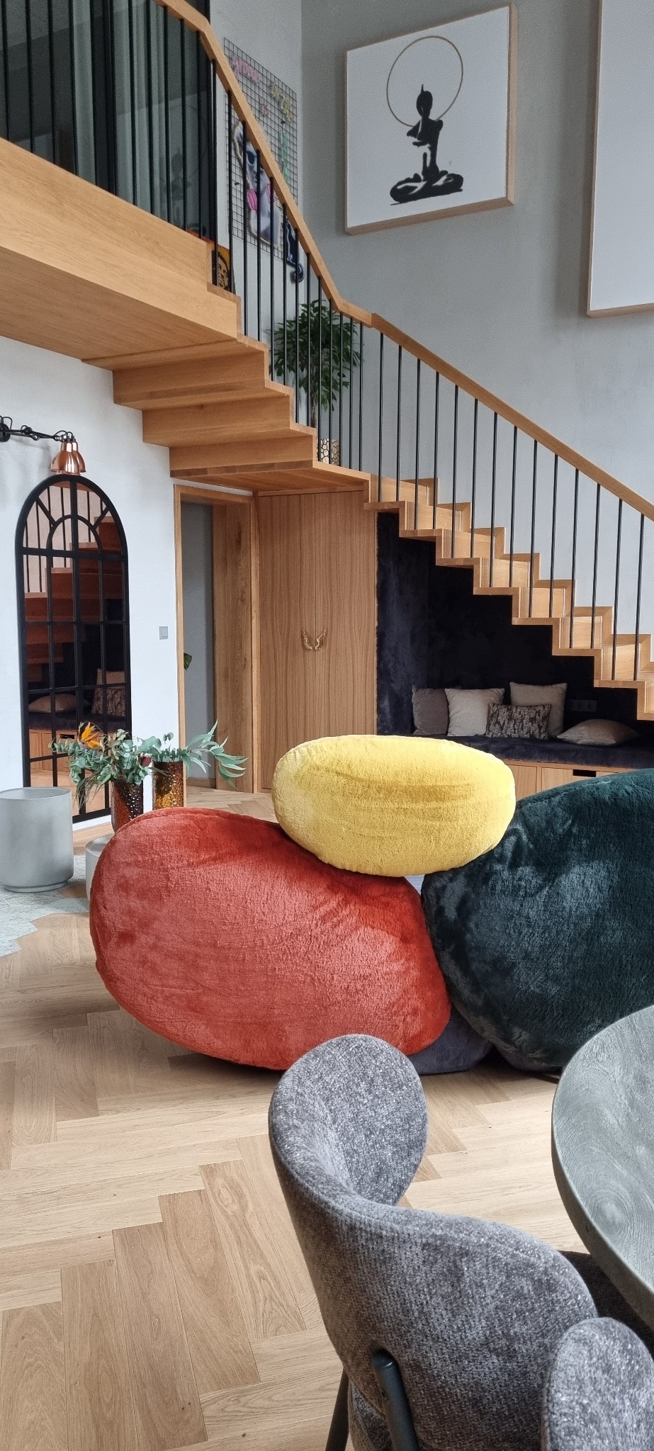 Wnętrze domu z drewnianymi schodami, dekoracyjnymi pufami w kształcie kamieni w kolorach żółtym, czerwonym i zielonym, krzesłem z szarej tkaniny i lustrem w stylu loft.