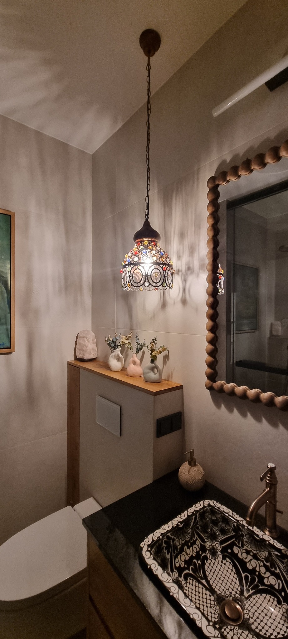 Wykończona łazienka z wiszącą, zdobioną lampą, umywalką w stylu retro z czarnym blatem i toaletą w tle, uzupełniona o dekoracje i lustro z drewnianą ramą.