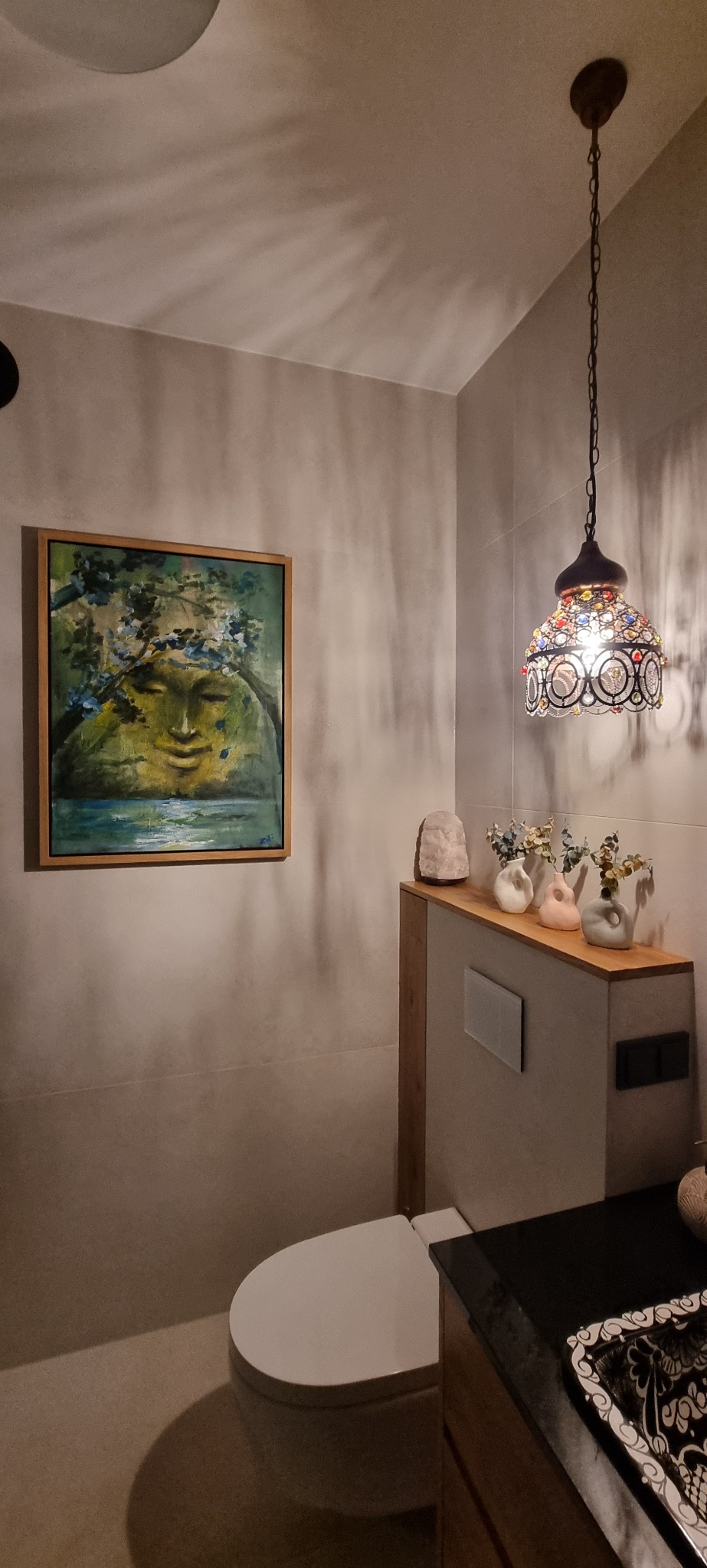 Umeblowana łazienka z toaletą, umywalką z czarnym blatem, obrazem z motywem twarzy i dekoracyjną lampą wiszącą z kolorowymi elementami.