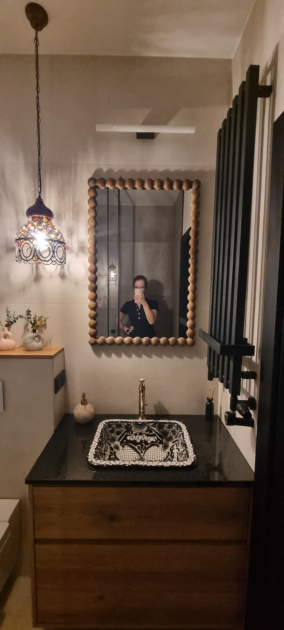 Umywalka nablatowa w czarno-białe wzory, rama lustra z drewnianymi kulkami, ozdobna lampa wisząca i grzejnik drabinkowy w łazience z beżowymi ścianami.