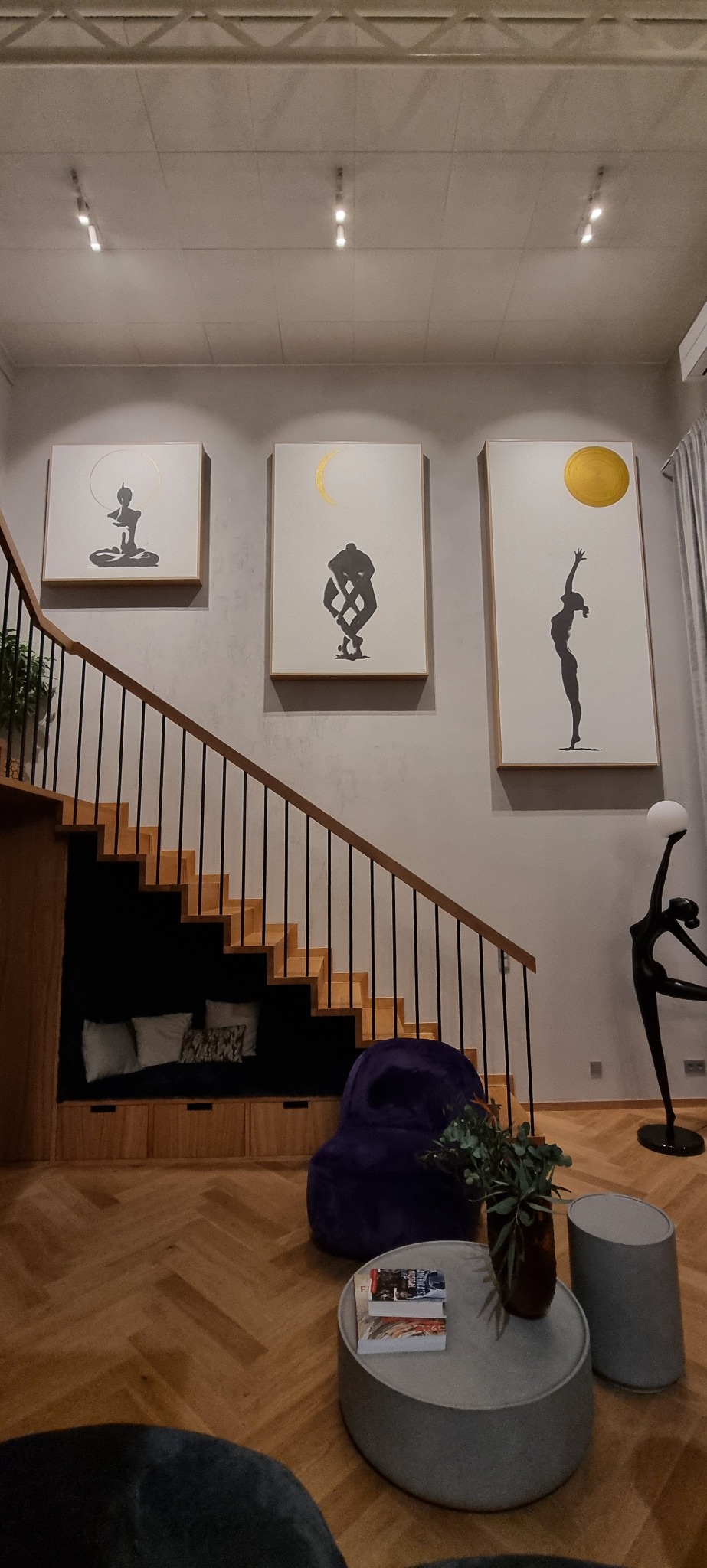 Salon z drewnianymi schodami, pod którymi znajduje się wnęka z siedziskiem i poduszkami, na ścianie wiszą trzy obrazy z motywami sylwetek i symboli słońca i księżyca, na podłodze parkiet w jodełkę.