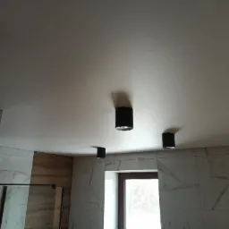 Biały, matowy sufit napinany z czterema czarnymi lampami punktowymi w łazience wykończonej płytkami imitującymi marmur i drewno.