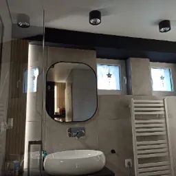 Nowoczesna łazienka z umywalką nablatową, lustrem w czarnej ramie, grzejnikiem drabinkowym i oświetleniem punktowym, wykończona płytkami w odcieniach szarości.