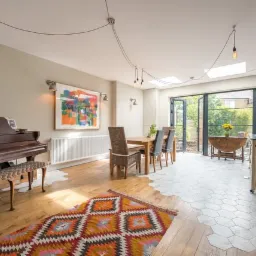 Salon z eklektyczną podłogą łączącą drewniane deski i heksagonalne płytki, pianinem, dywanem w etniczne wzory i otwartym przejściem do ogrodu przez składane drzwi.