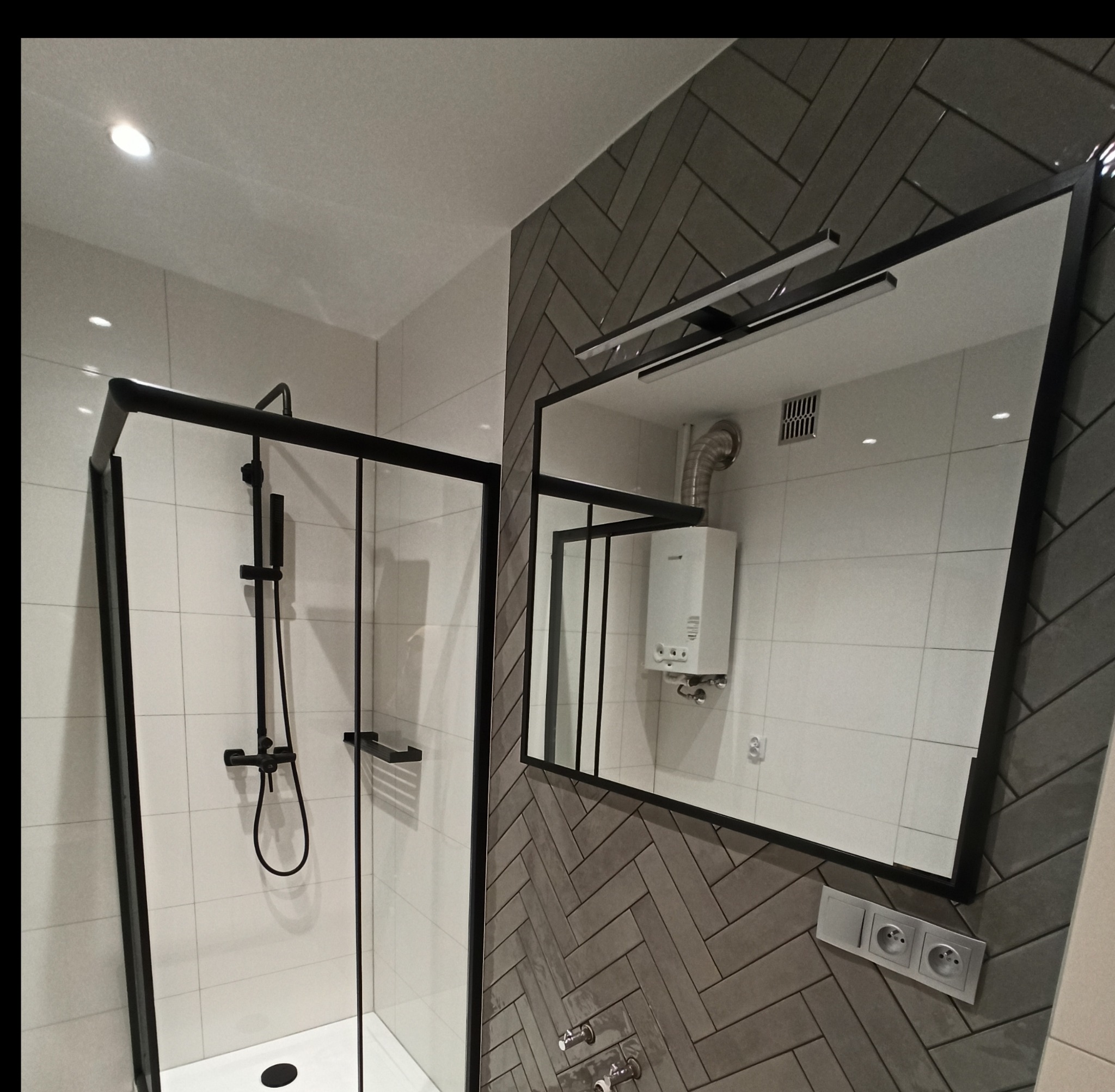 Nowoczesna łazienka z kabiną prysznicową typu walk-in z czarnymi ramami, lustrem z oświetleniem i ścianą wyłożoną szarą glazurą w jodełkę.