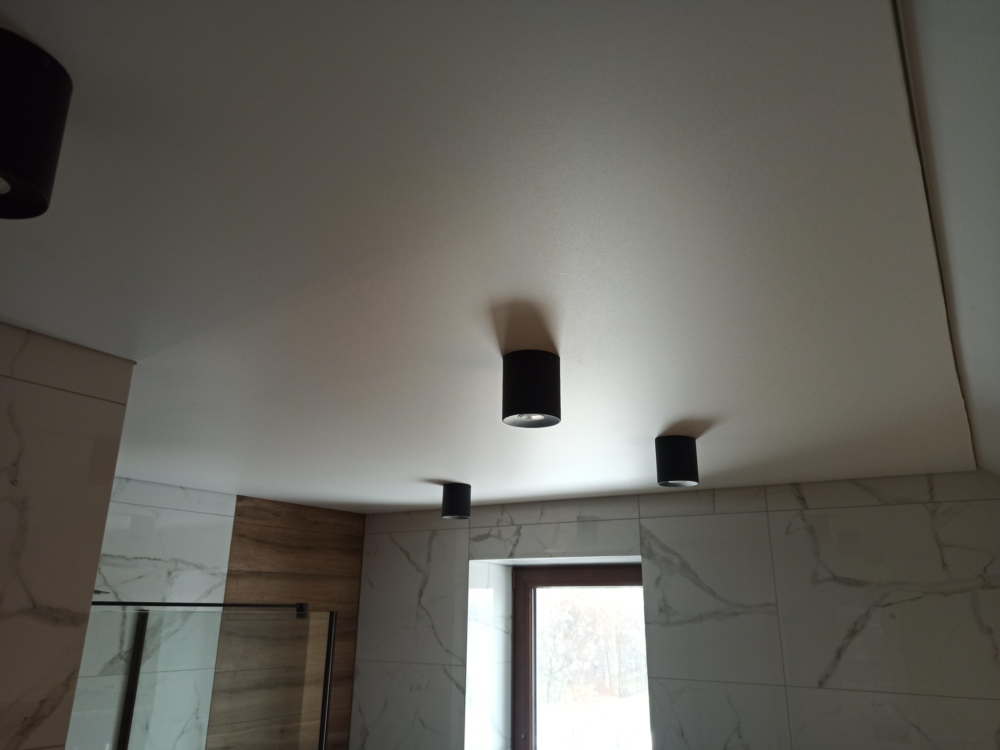 Biały, matowy sufit napinany z czterema czarnymi lampami punktowymi w łazience wykończonej płytkami imitującymi marmur i drewno.