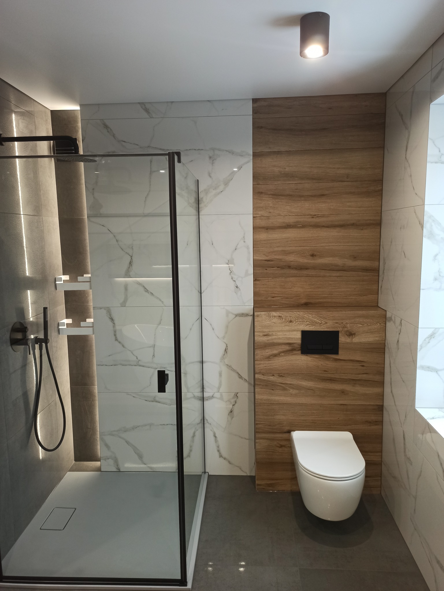 Nowoczesna łazienka z kabiną prysznicową typu walk-in, płytkami imitującymi marmur, drewnianą okładziną ścienną za toaletą podtynkową oraz szarą podłogą.