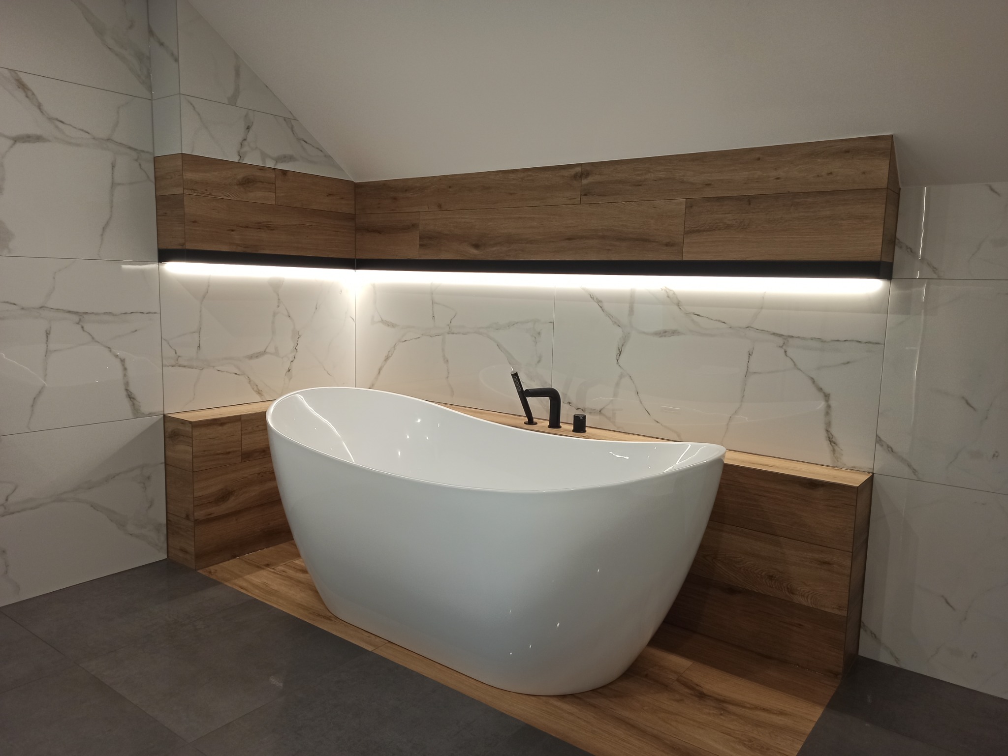Nowoczesna łazienka z wolnostojącą białą wanną, czarną armaturą i marmurowymi płytkami na ścianach, podświetlona listwa LED nad wanną.
