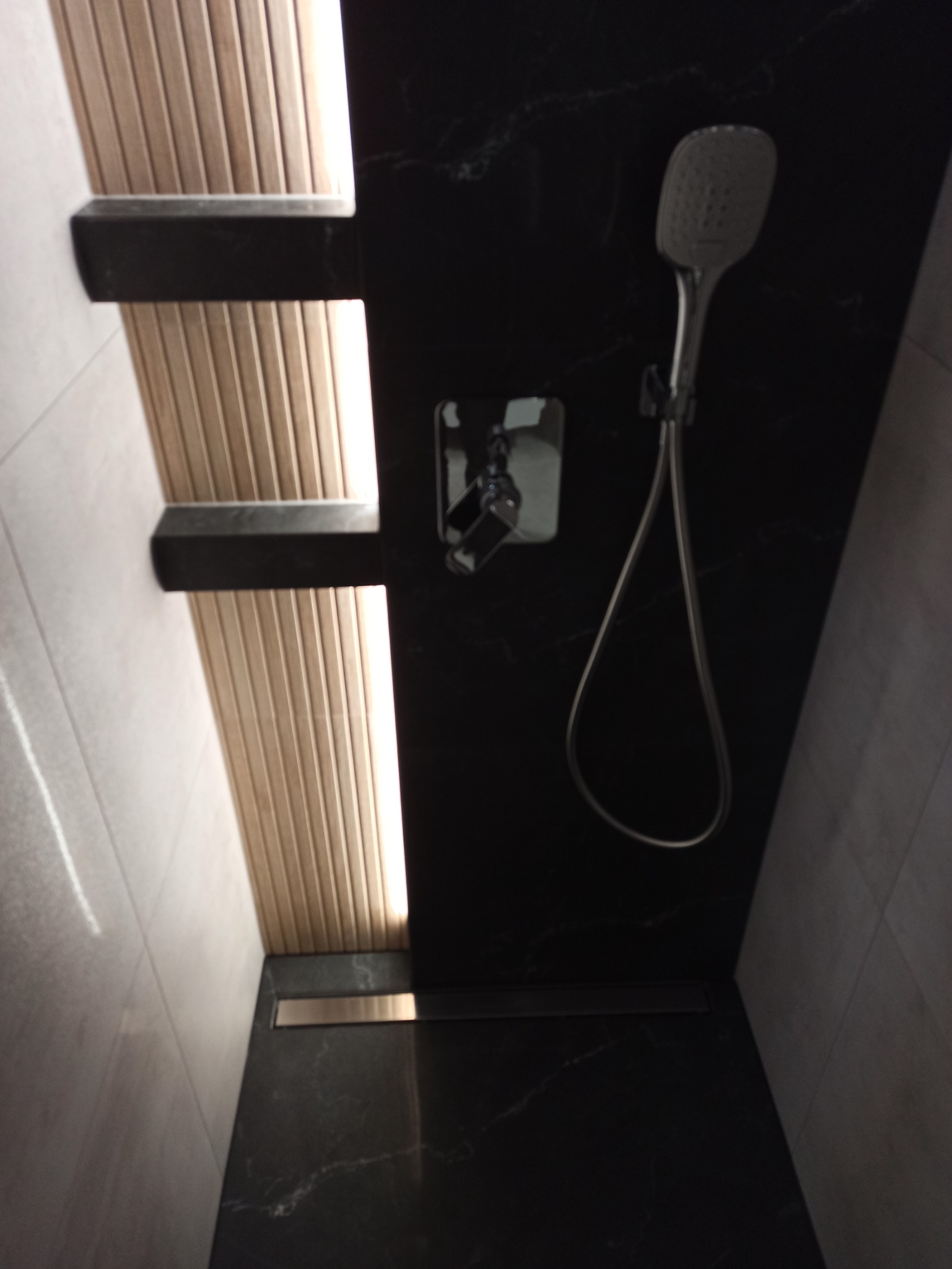 Nowoczesna łazienka z czarną marmurową ścianą, deszczownicą, ukrytym odpływem liniowym i drewnianymi panelami z podświetleniem LED.