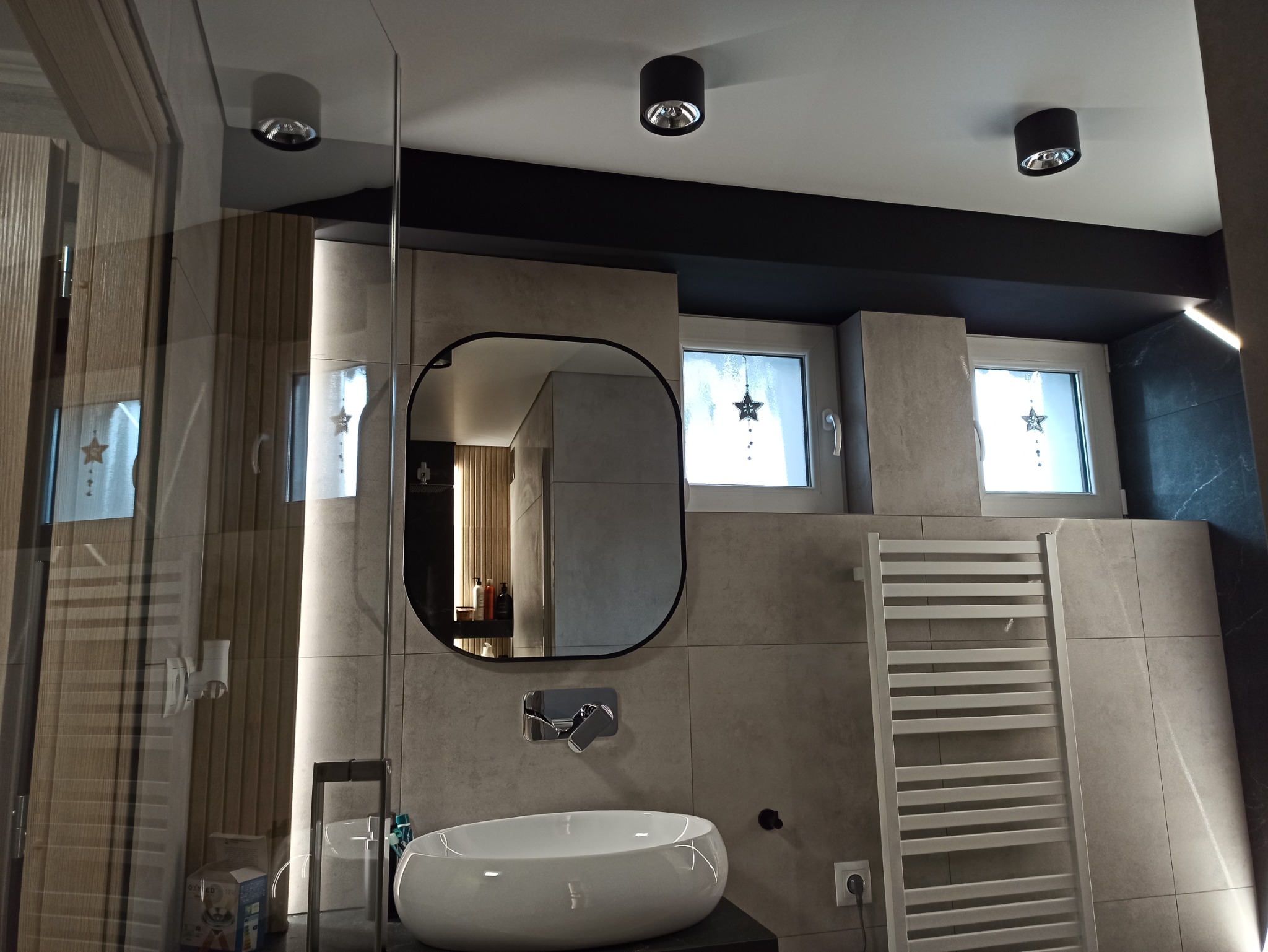 Nowoczesna łazienka z umywalką nablatową, lustrem w czarnej ramie, grzejnikiem drabinkowym i oświetleniem punktowym, wykończona płytkami w odcieniach szarości.