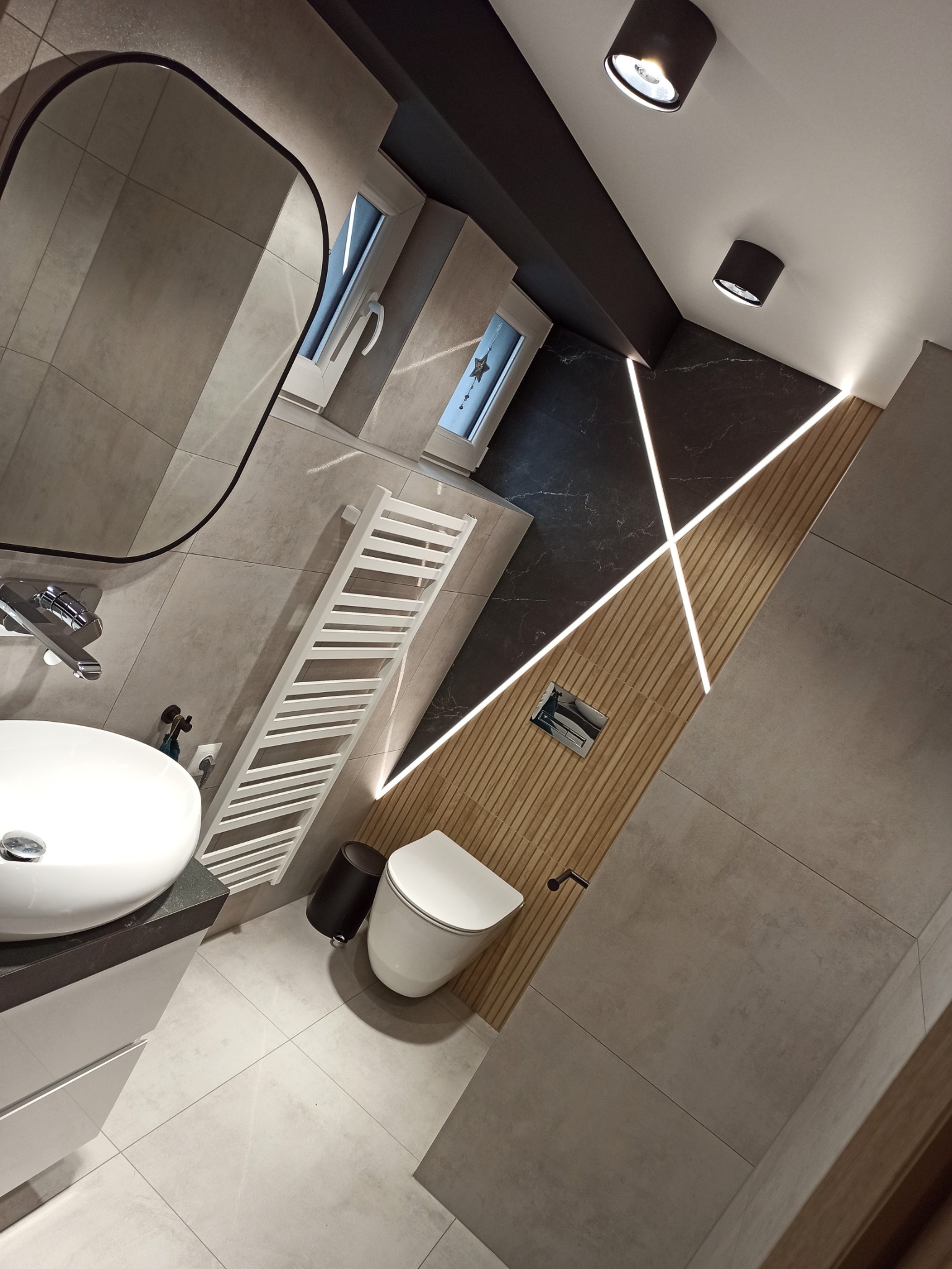 Nowoczesna łazienka z szarymi płytkami, drewnianą ścianą z podświetleniem LED w kształcie krzyża, białą umywalką nablatową i grzejnikiem drabinkowym.