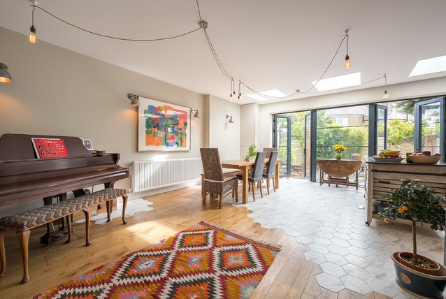 Salon z eklektyczną podłogą łączącą drewniane deski i heksagonalne płytki, pianinem, dywanem w etniczne wzory i otwartym przejściem do ogrodu przez składane drzwi.