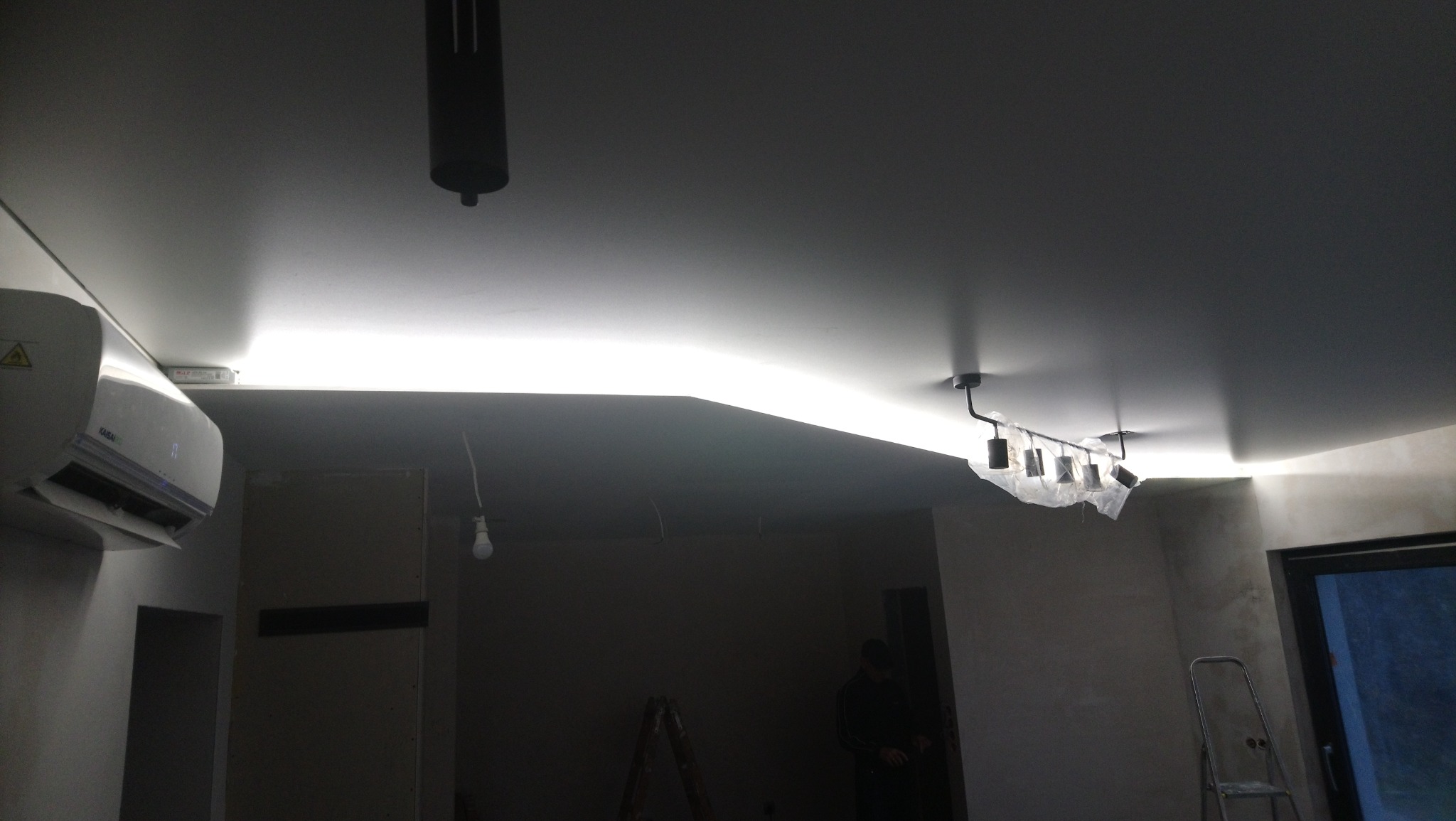 Częściowo wykończone wnętrze z podwieszonym sufitem z oświetleniem LED, klimatyzacją na ścianie, lampą sufitową zabezpieczoną folią, drabina i osoba w tle.