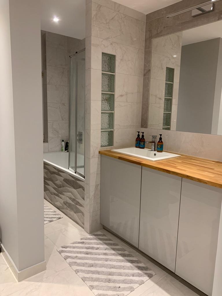 Nowoczesna łazienka z marmurowymi płytkami, drewnianym blatem, szarymi szafkami pod umywalką i szklanymi luksferami w ścianie, uzupełniona o pasiaste dywaniki.