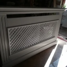 Biała, dekoracyjna osłona grzejnika z kratką w romby, widoczna w warsztacie stolarskim, z elementami dekoracyjnymi na górze i po bokach.