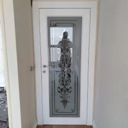 Białe drzwi wewnętrzne z ozdobnym, piaskowanym wzorem na szkle, widoczne z bliska na tle jasnej ściany i podłogi imitującej drewno.