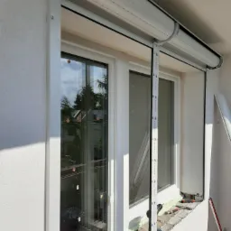 Biała roleta zewnętrzna częściowo opuszczona, widoczne okno z ramą PCV, narzędzia i pianka montażowa na parapecie, etap instalacji.