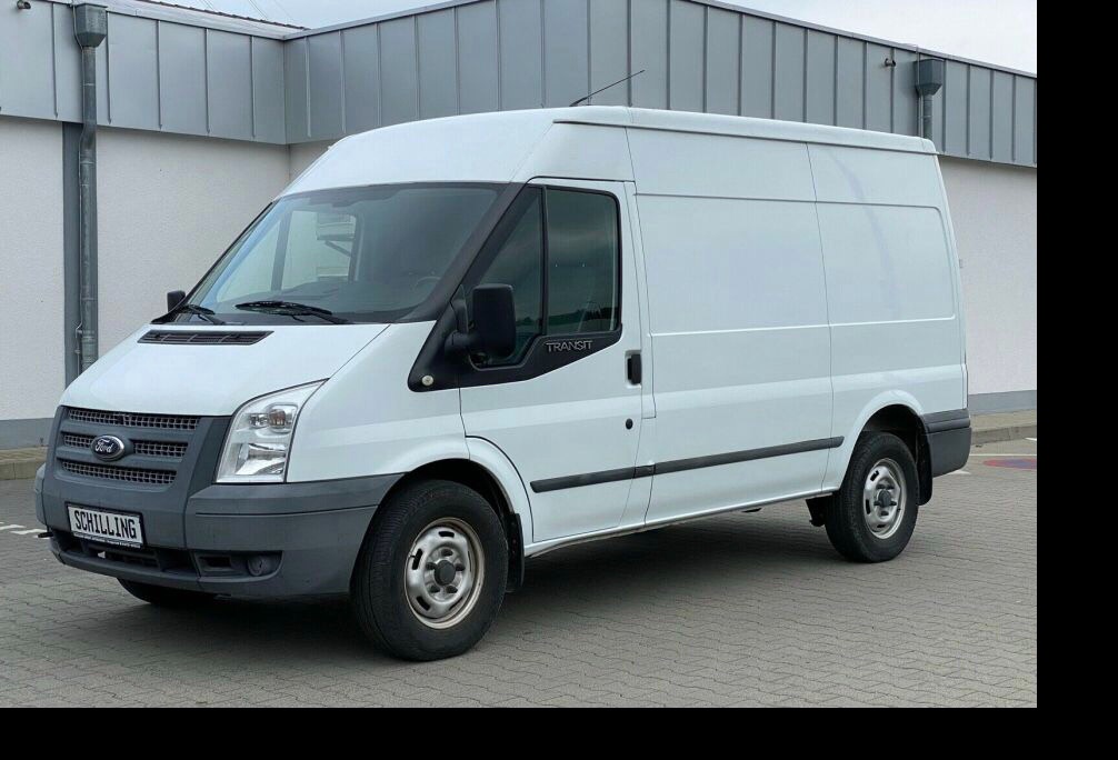 Biały Ford Transit z widoczną tablicą rejestracyjną SCHILLING, zaparkowany na szarym bruku przed budynkiem z szarą elewacją.