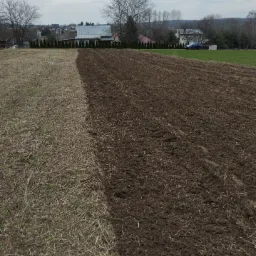 Częściowo niwelowany teren z widoczną granicą między obszarem z suchą trawą a świeżo przekopaną ziemią, w tle zabudowania wiejskie i drzewa.
