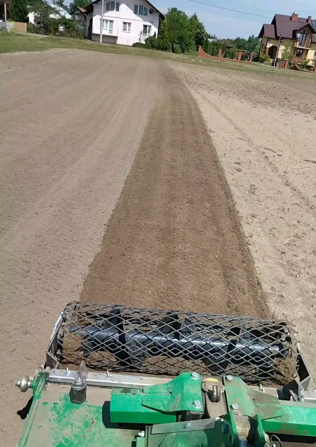 Widok z perspektywy operatora na walec ugniatający świeżo wyrównany teren pod trawnik, widoczne domy w tle.