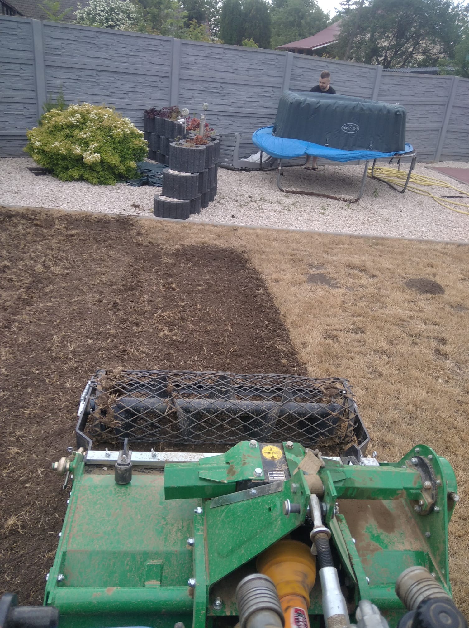 Przygotowanie gleby pod trawnik za pomocą specjalistycznej maszyny, widoczne świeżo spulchniona ziemia i fragment trawnika przed zabiegiem, w tle ogrodzenie i osoba przy jacuzzi.