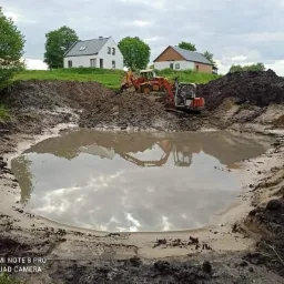 Dwa urządzenia budowlane, koparka i spycharka, pracują przy wykopie dużego, owalnego zbiornika wodnego, w którym odbija się pochmurne niebo. W tle widoczne dwa budynki mieszkalne na wzgórzu.