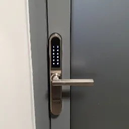 Szare drzwi z zamontowanym srebrnym zamkiem elektronicznym Smart Lock z klawiaturą numeryczną i klamką.