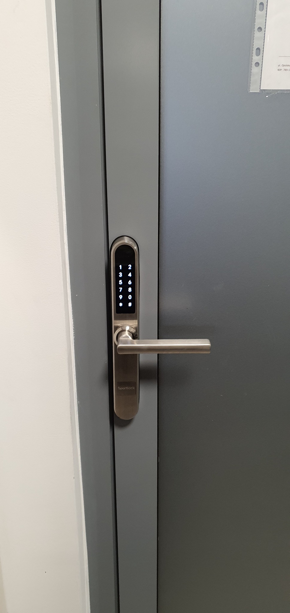 Szare drzwi z zamontowanym srebrnym zamkiem elektronicznym Smart Lock z klawiaturą numeryczną i klamką.