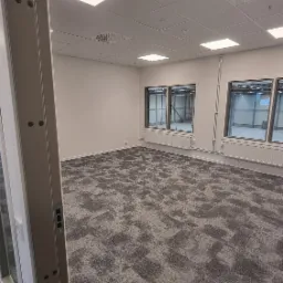 Puste biuro z szarą wykładziną dywanową w kratkę, białymi ścianami i oknami z widokiem na halę magazynową, widoczne elementy systemu wentylacji na suficie.