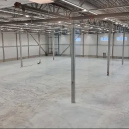 Przestronne wnętrze hali magazynowej z betonową posadzką, widoczne stalowe słupy podtrzymujące konstrukcję dachu, liczne świetlówki oraz instalacje pod sufitem, białe ściany z oknami i drzwiami...