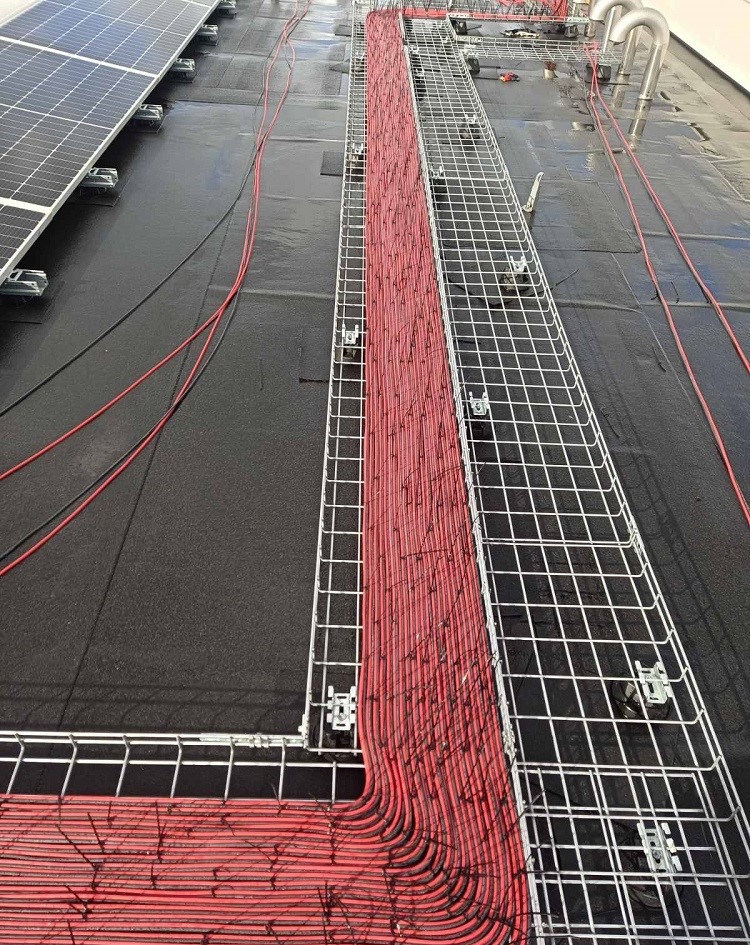 Widok z góry na dach z panelami słonecznymi i korytem kablowym wypełnionym dużą ilością czerwonych kabli, zabezpieczonych metalową siatką.