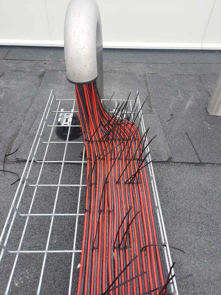 Kable elektryczne czerwono-czarne ułożone w metalowej korycie kablowej, zabezpieczone czarnymi opaskami zaciskowymi, wychodzące z metalowej rury na dachu.