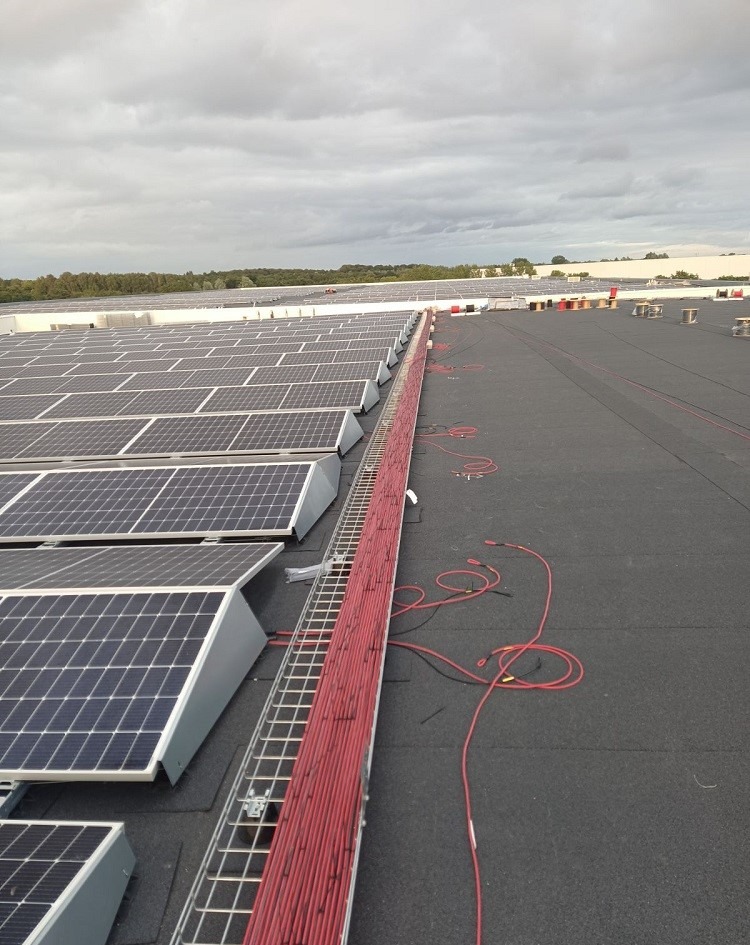 Instalacja paneli fotowoltaicznych na dachu budynku przemysłowego, widoczne koryto kablowe wypełnione czerwonymi przewodami, w tle liczne panele i pochmurne niebo.