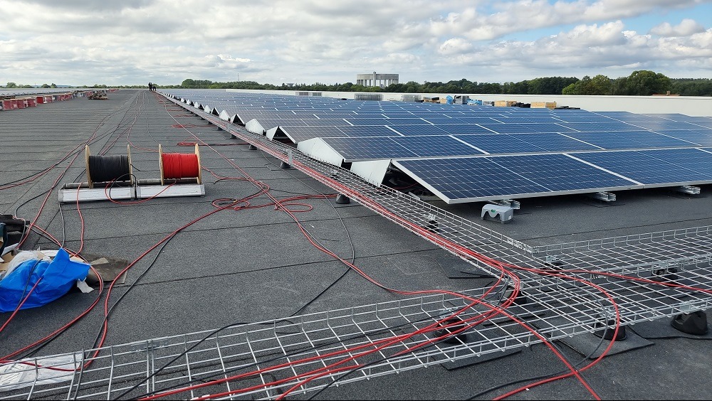 Instalacja paneli słonecznych na dachu budynku, widoczne kable prowadzone w metalowych korytach, dwie szpule z czerwonym i czarnym przewodem, w tle inne panele i budynek.