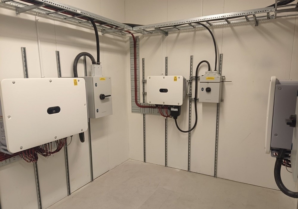 Wnętrze pomieszczenia z zamontowanymi białymi falownikami i skrzynkami elektrycznymi, kable prowadzone w korytach na suficie i po ścianach.