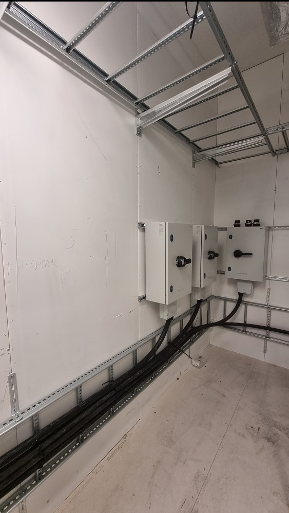 Wnętrze pomieszczenia z trzema białymi skrzynkami elektrycznymi zamontowanymi na ścianie, kable prowadzone w metalowych korytach, widoczne metalowe profile na ścianach i suficie.