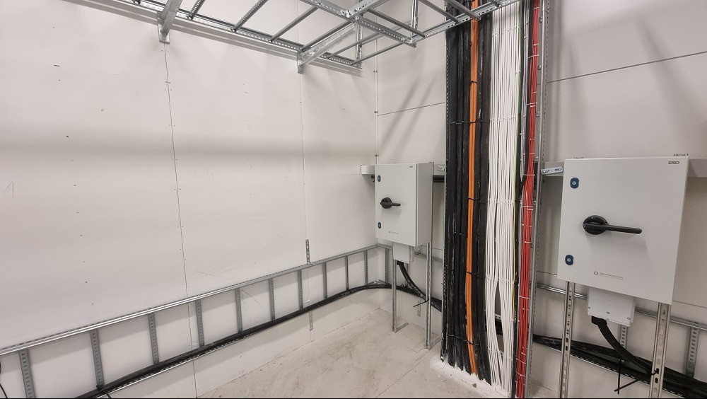 Wnętrze pomieszczenia technicznego z instalacją elektryczną: białe ściany, metalowe profile pod sufitem i na ścianach, pionowe wiązki kabli w kolorach czarnym, białym i czerwonym, dwie białe...