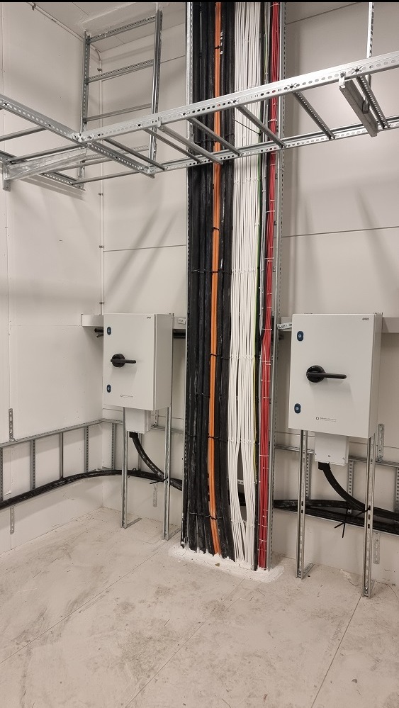Rząd kabli elektrycznych w korytach, zabezpieczonych przy podłodze białą masą uszczelniającą, z dwoma skrzynkami sterowniczymi po bokach, zamontowanymi na metalowych wspornikach.