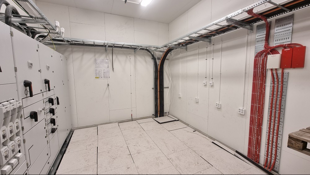 Wnętrze pomieszczenia technicznego z rozdzielnicą elektryczną, korytkami kablowymi i przewodami ułożonymi wzdłuż ścian.