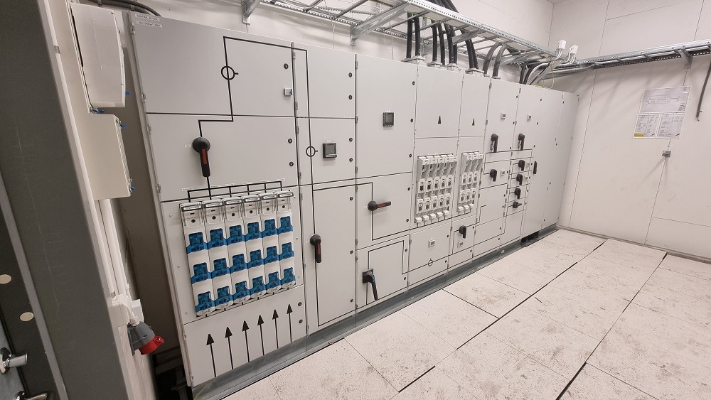 Rozbudowana szafa elektryczna z licznymi zabezpieczeniami i schematami, widoczne kable w korytach kablowych pod sufitem, na ścianie instrukcja obsługi.