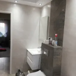 Nowoczesna łazienka w Tenczynku z szarymi płytkami imitującymi kamień, podwieszana umywalka z szafką, toaleta, na półce środki czystości, widoczny odkurzacz na podłodze.