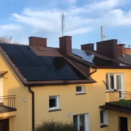 Żółty dom jednorodzinny z brązowym dachem, na którym zamontowane są czarne panele słoneczne. Na dachu widoczna antena telewizyjna oraz komin. Częściowo widoczne inne budynki w tle.