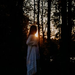 Foto-Pocisk - Sylwetka dziewczynki w lesie, podświetlona zachodzącym słońcem. Dziewczynka ubrana w biały sweter z frędzlami, stoi tyłem do obiektywu, patrząc w dal.