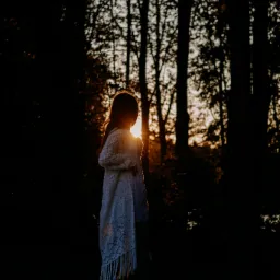 Sylwetka dziewczynki w lesie, podświetlona zachodzącym słońcem. Dziewczynka ubrana w biały sweter z frędzlami, stoi tyłem do obiektywu, patrząc w dal.